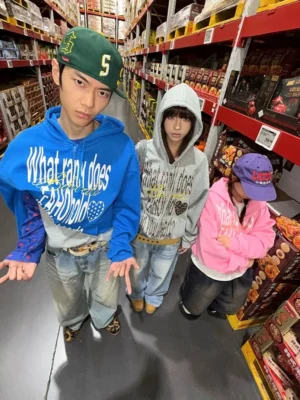 Blue graphic zip-up hoodie with bold white front lettering styled on three models in a colorful supermarket snack aisle