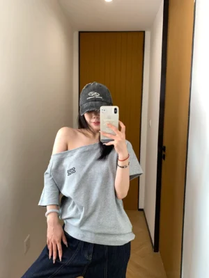 Side mirror view showing the loose fit and one-shoulder neckline of a gray Korean streetwear logo T-shirt with dark denim