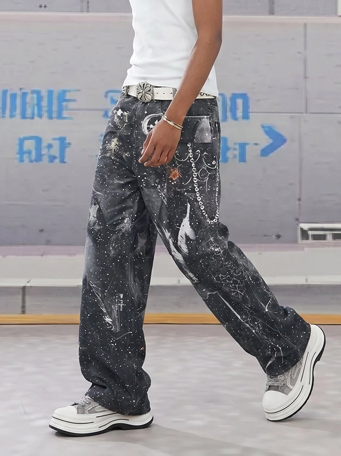 Male streetwear outfit styled with gray frayed canvas platform sneakers, wide-leg splatter print jeans, and layered chain accessories