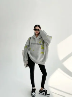 Full body model wearing a gray oversized graphic hoodie with lime green splatter details, black leggings, and sneakers