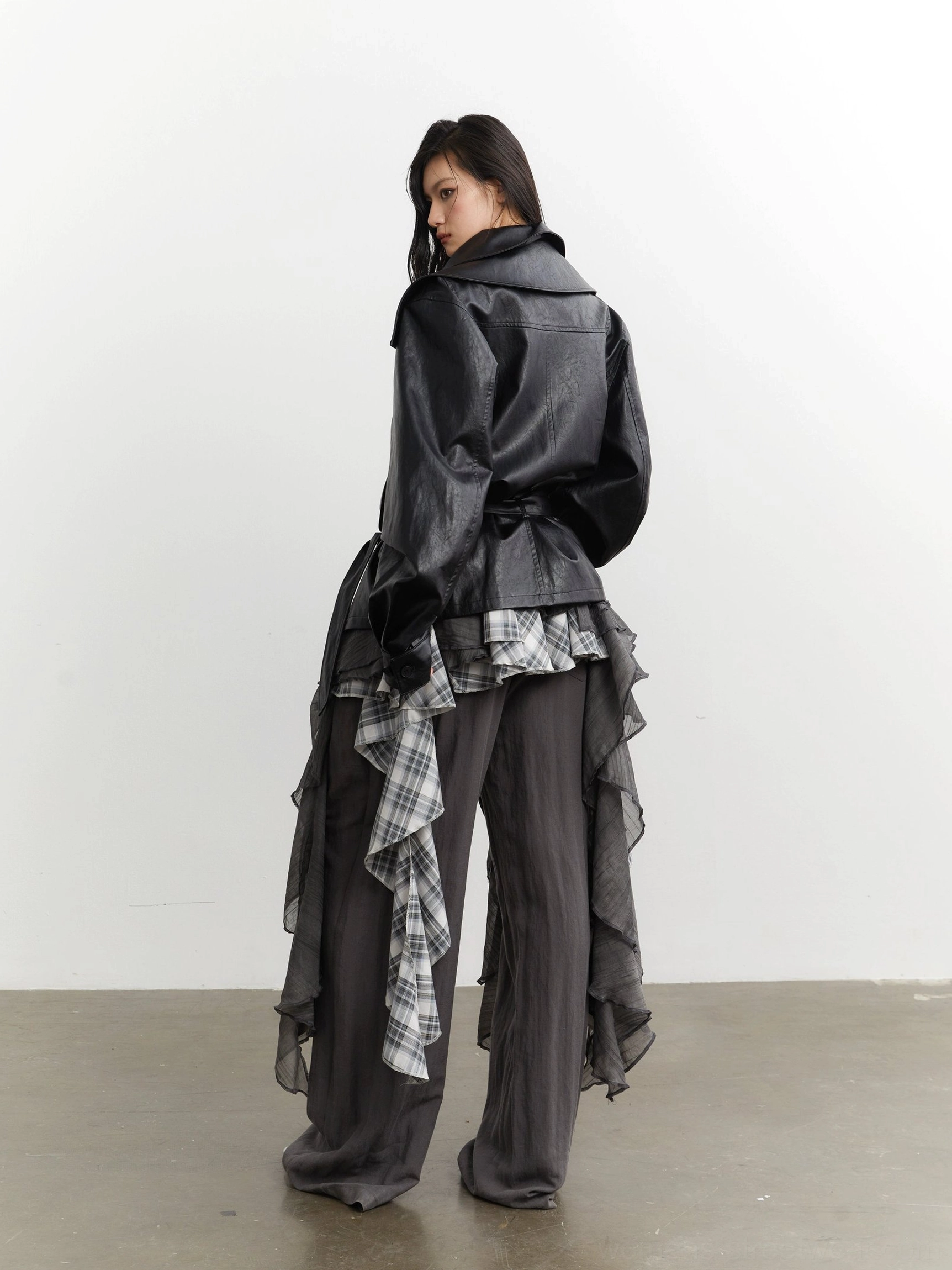 Back view of dark layered streetwear outfit styled with gray plaid ruffle detail and black belted leather jacket