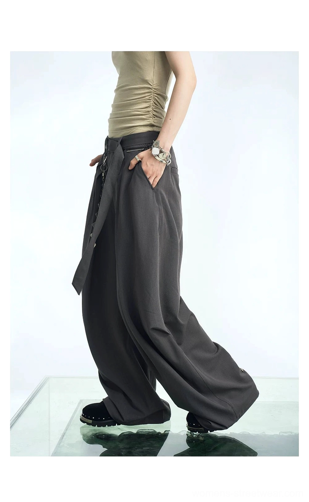Dark gray pleated balloon wide-leg streetwear pants shown in a walking side view with high waist, fluid drape, cropped beige top, and casual sneakers