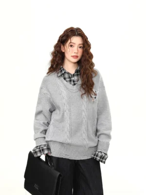 Front view of a women gray preppy cable knit V-neck sweater with plaid faux shirt collar, exposed cuffs, embroidered crest badge, wide black trousers, and structured briefcase
