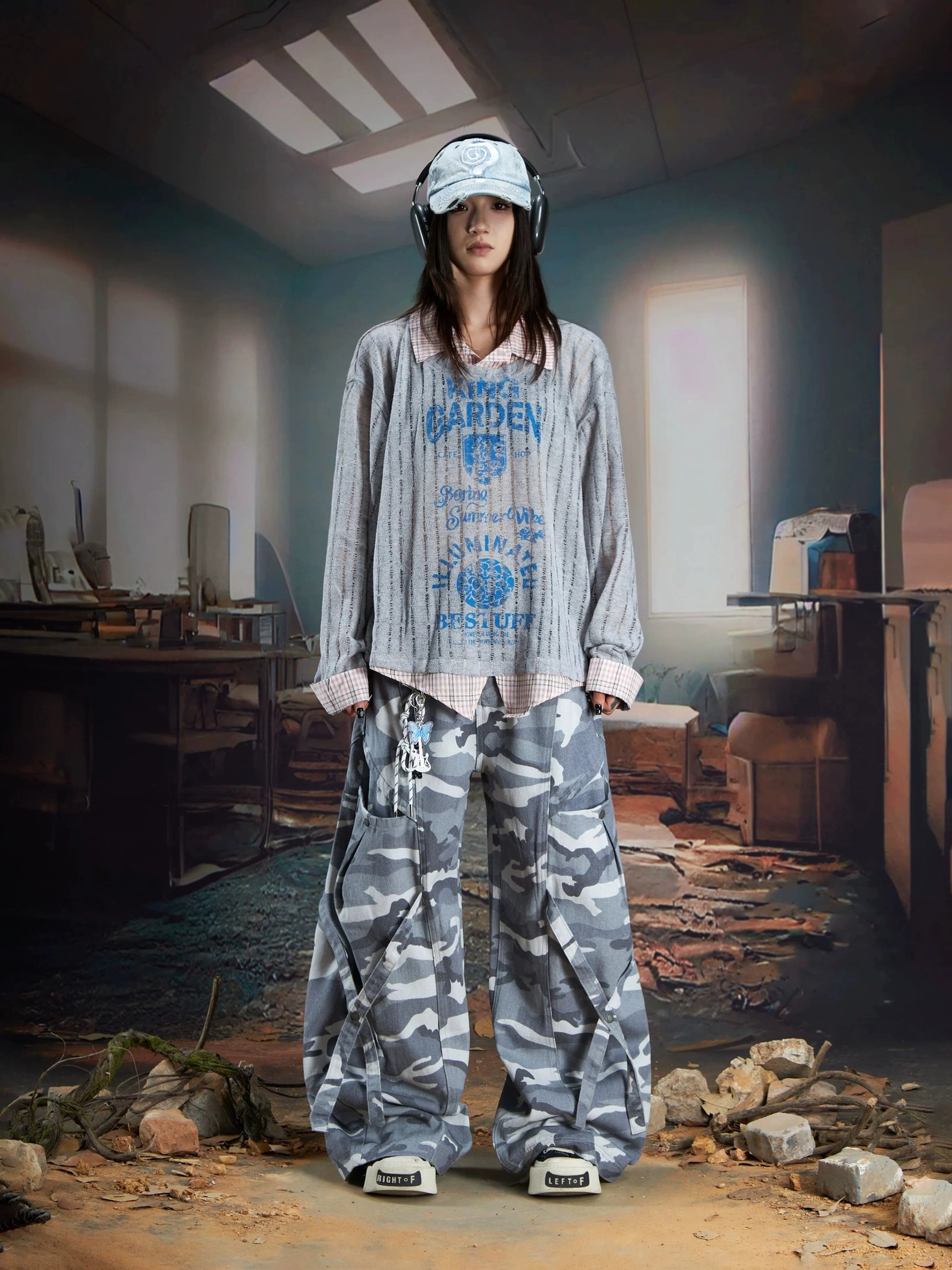 Female model in a gray ribbed sheer layered shirt sweater with blue retro graphics, contrast collar details, cap, headphones, and camouflage cargo pants