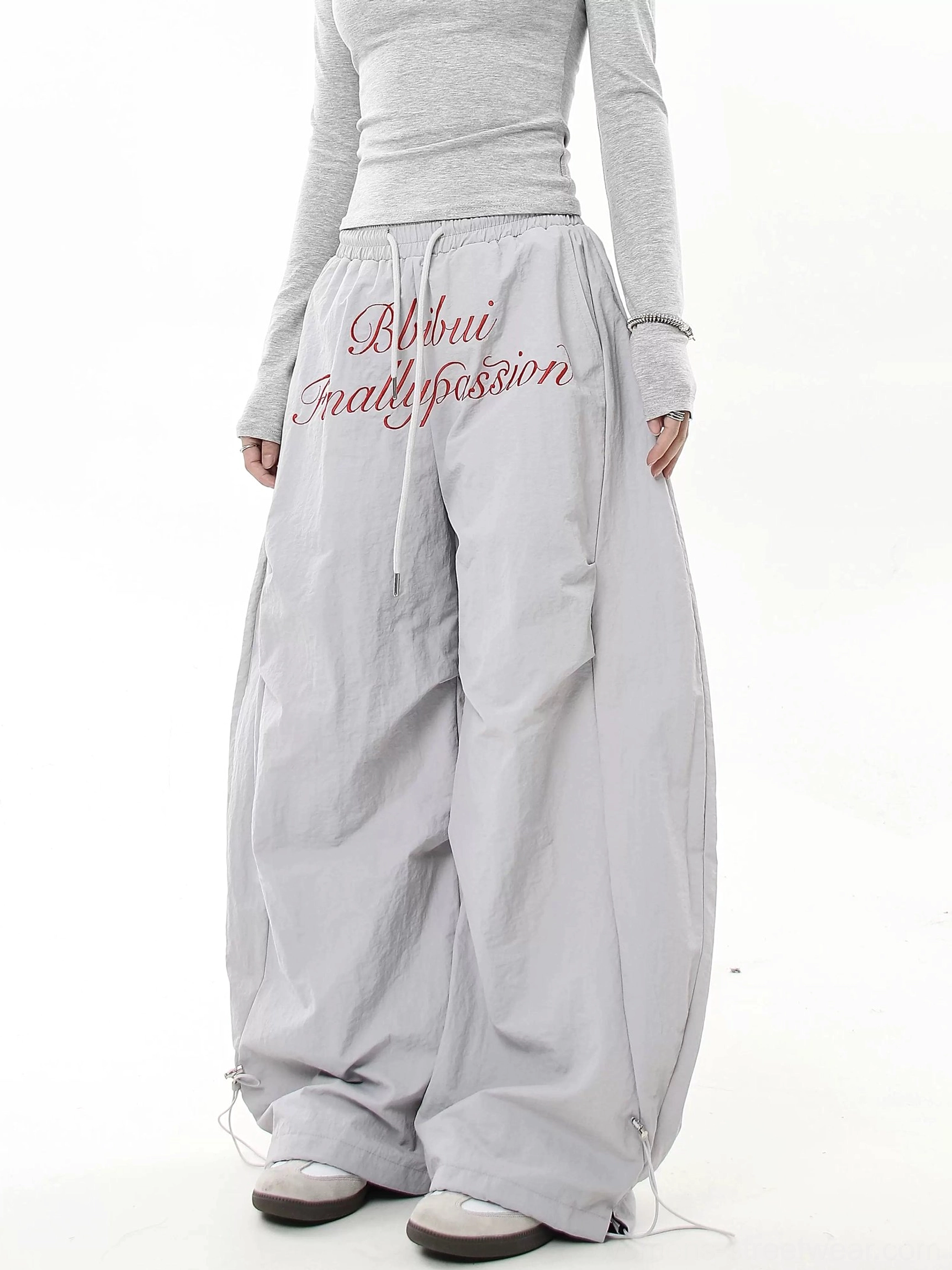 Light gray oversized streetwear parachute pants with red script detail, drawstring waist, loose wide legs and adjustable ankle cuffs