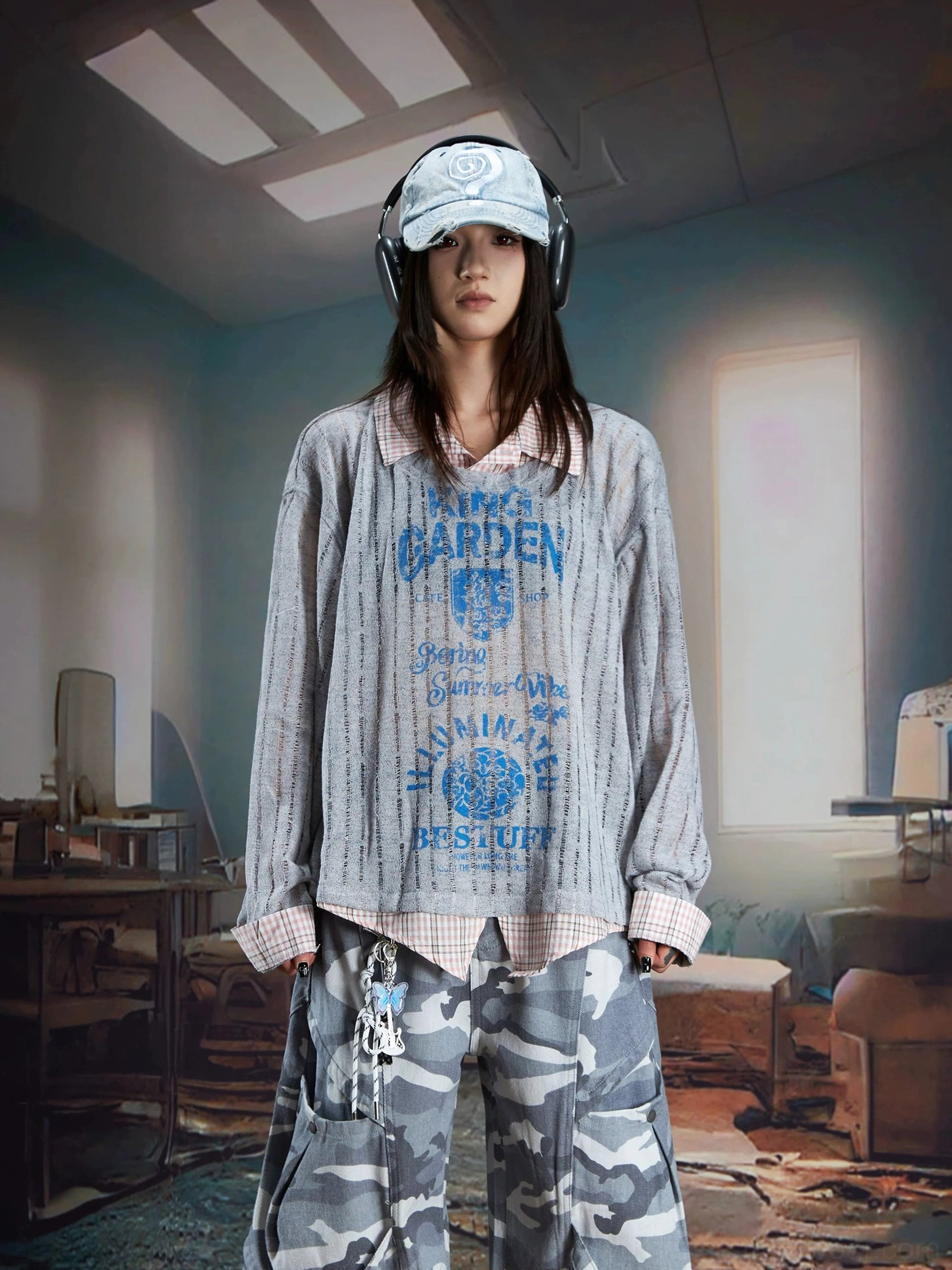 Female model wearing a gray sheer layered vintage print shirt sweater with blue KING GARDEN style graphics, headphones, cap, and gray camouflage cargo pants