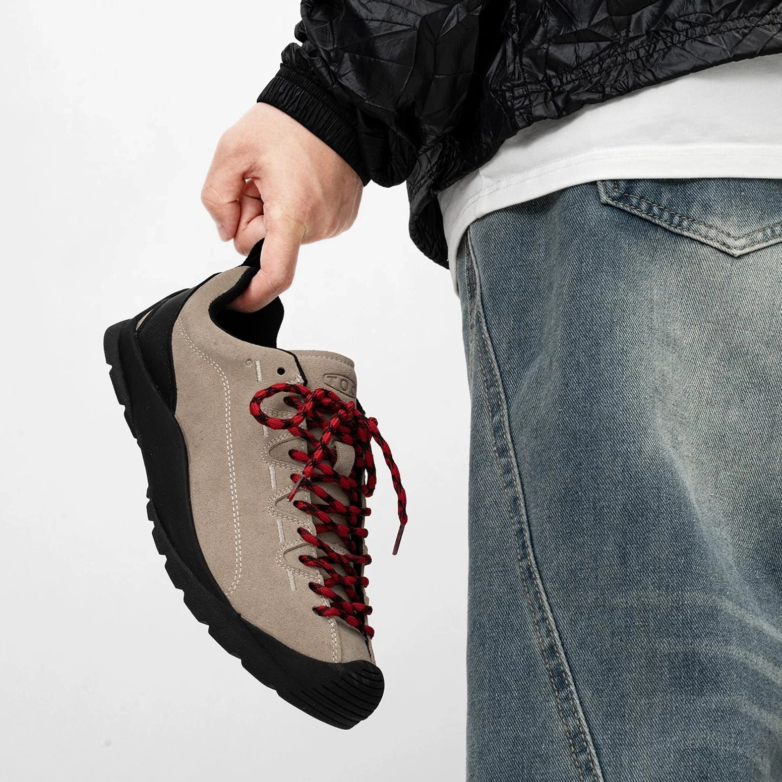 Gray taupe suede hiking sneakers with dark red woven laces, low-top collar and black rugged rubber outsole shown in hand with baggy denim
