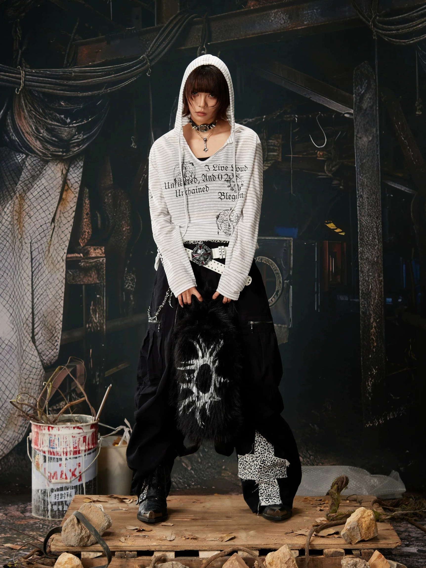Full-body view of a gray and white striped hooded long sleeve streetwear top styled with black cargo pants in an industrial setting