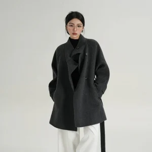 Three-quarter view of a women's grayish-gray wool coat featuring a high asymmetric funnel collar, double-breasted buttons, relaxed fit, side pockets, and back waist belt