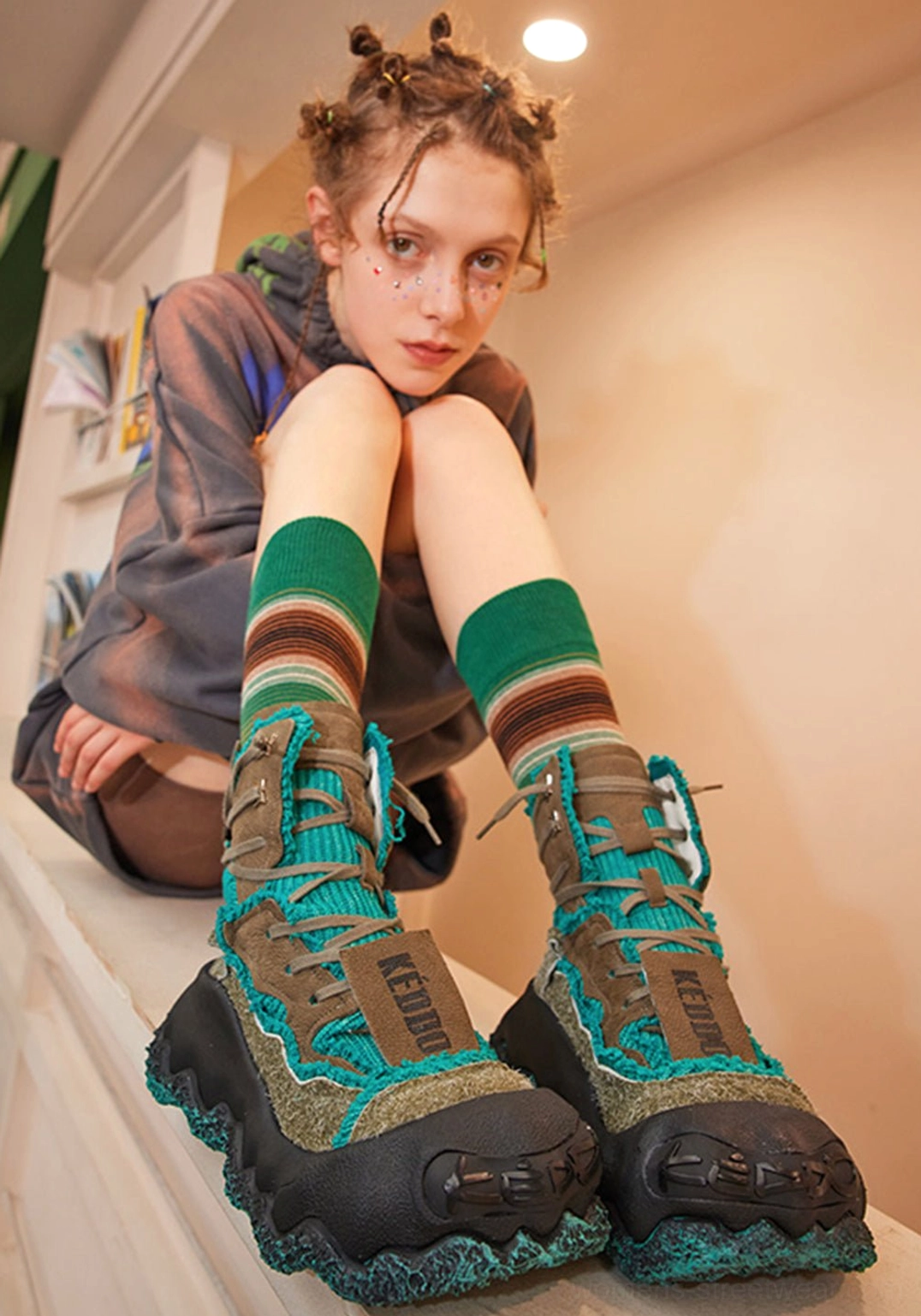 Female model wearing green abstract splash platform combat boots with striped teal socks in a relaxed indoor Y2K streetwear setting