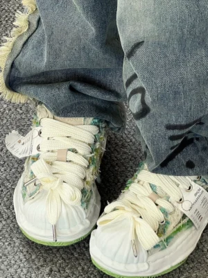 Close-up overhead view of green frayed textile streetwear sneakers worn with frayed hem denim, showing the textured tweed upper, oversized laces, shell toe, product tag, and layered platform sole