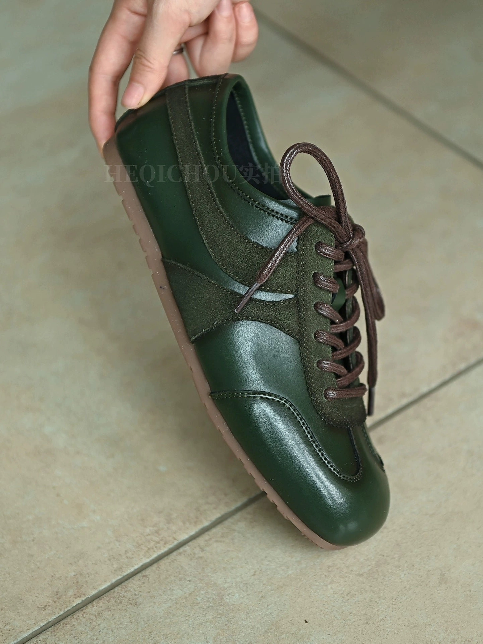 Dark green retro sneakers held above a tiled floor, showing mixed suede and leather panels, brown laces, and a classic low-top silhouette