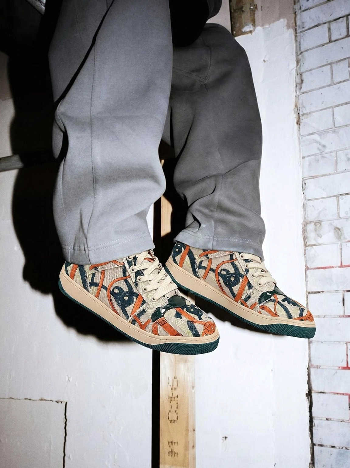 Overhead on-foot view of green sole graffiti canvas low-top sneakers styled with gray relaxed trousers by an industrial brick wall