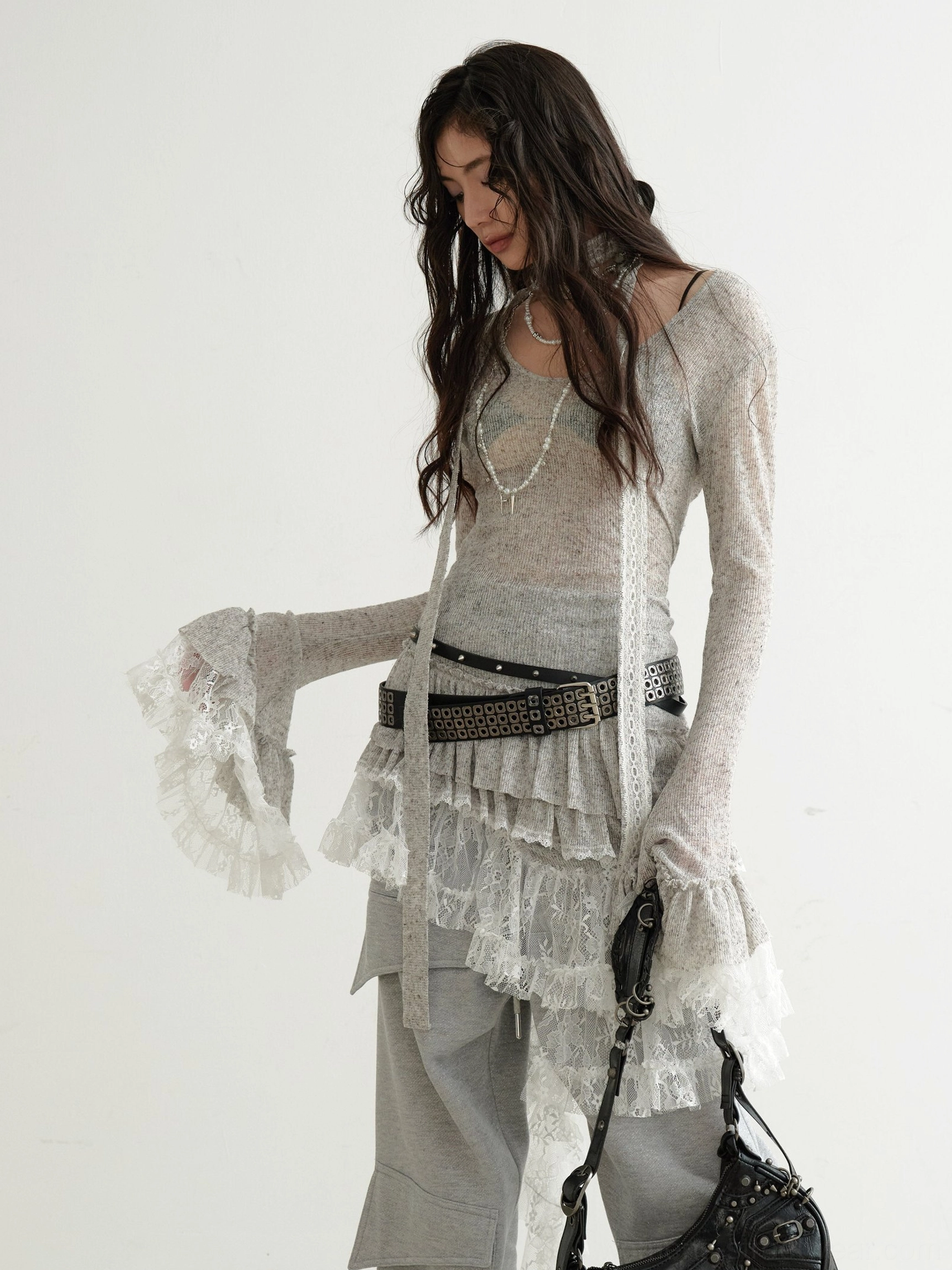 Side view of a woman wearing a grey asymmetrical lace knit dress with long slim sleeves, ruffled cuffs, layered necklaces, studded belt, and vintage shoulder bag