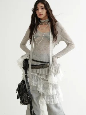 Front three-quarter view of a woman wearing a grey asymmetrical lace panel knit dress with layered ruffle hem, wide-leg trousers, studded belt, and vintage shoulder bag