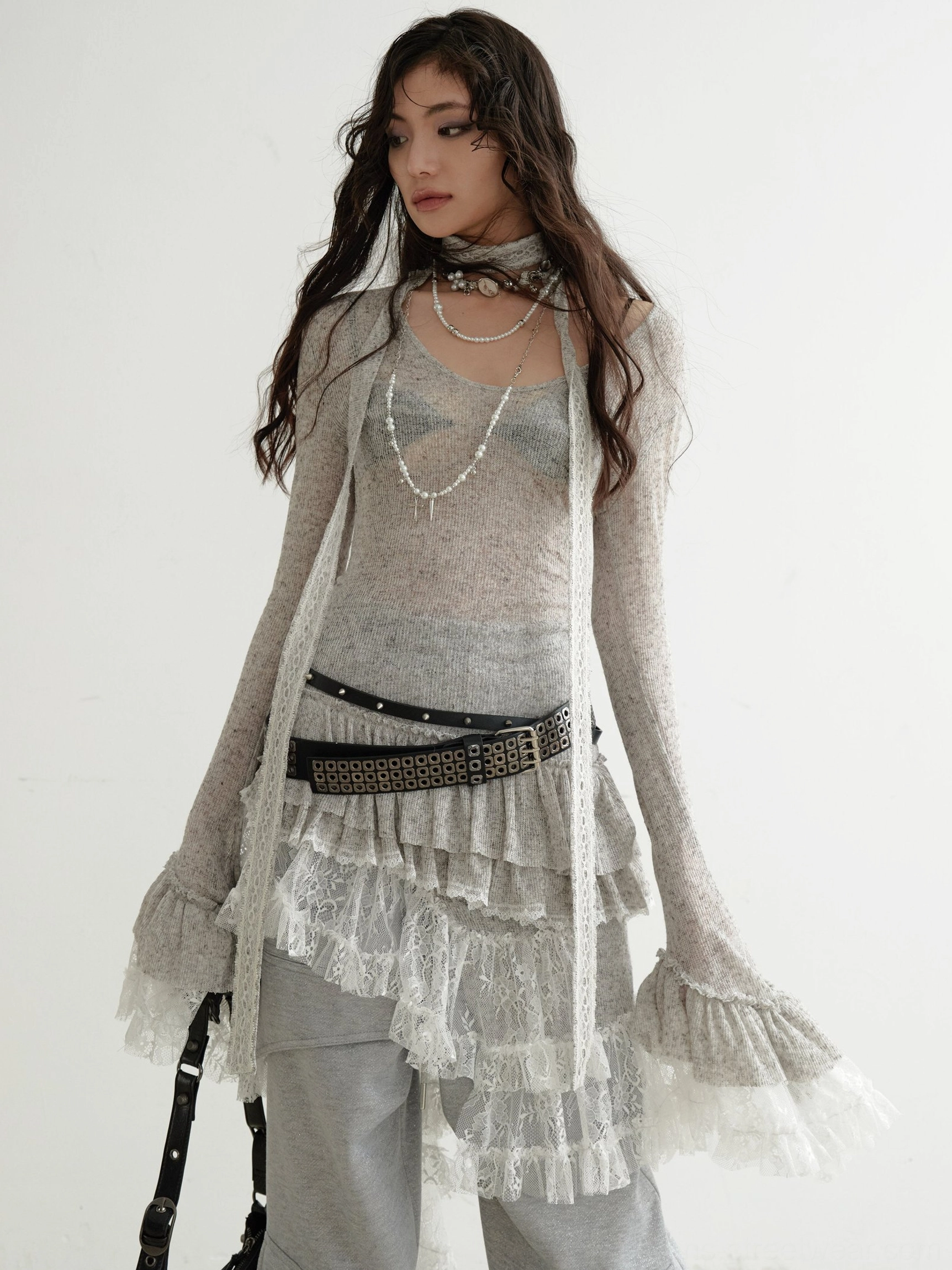 Side front view of grey lace panel knit dress with bell-shaped ruffled cuffs, layered asymmetrical lace skirt, wide-leg trousers, studded belt, and shoulder bag