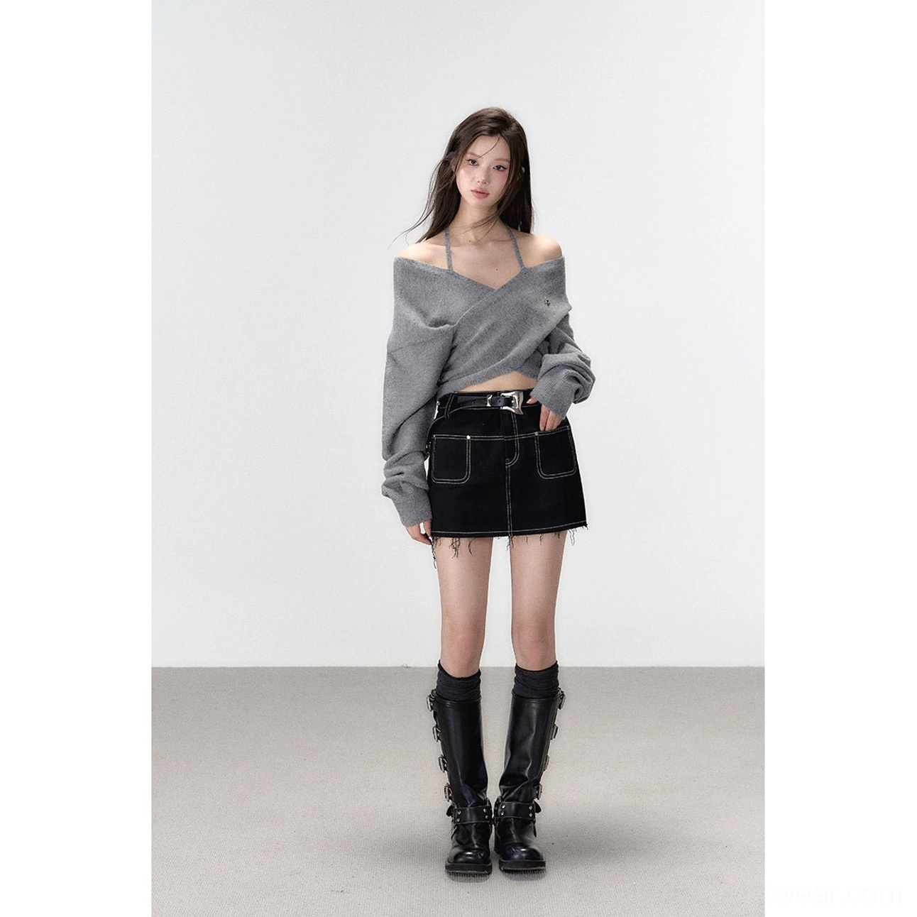 Full standing view of a model wearing a grey long-sleeve off-shoulder halter knit top with a cropped hem, denim skirt, and boots