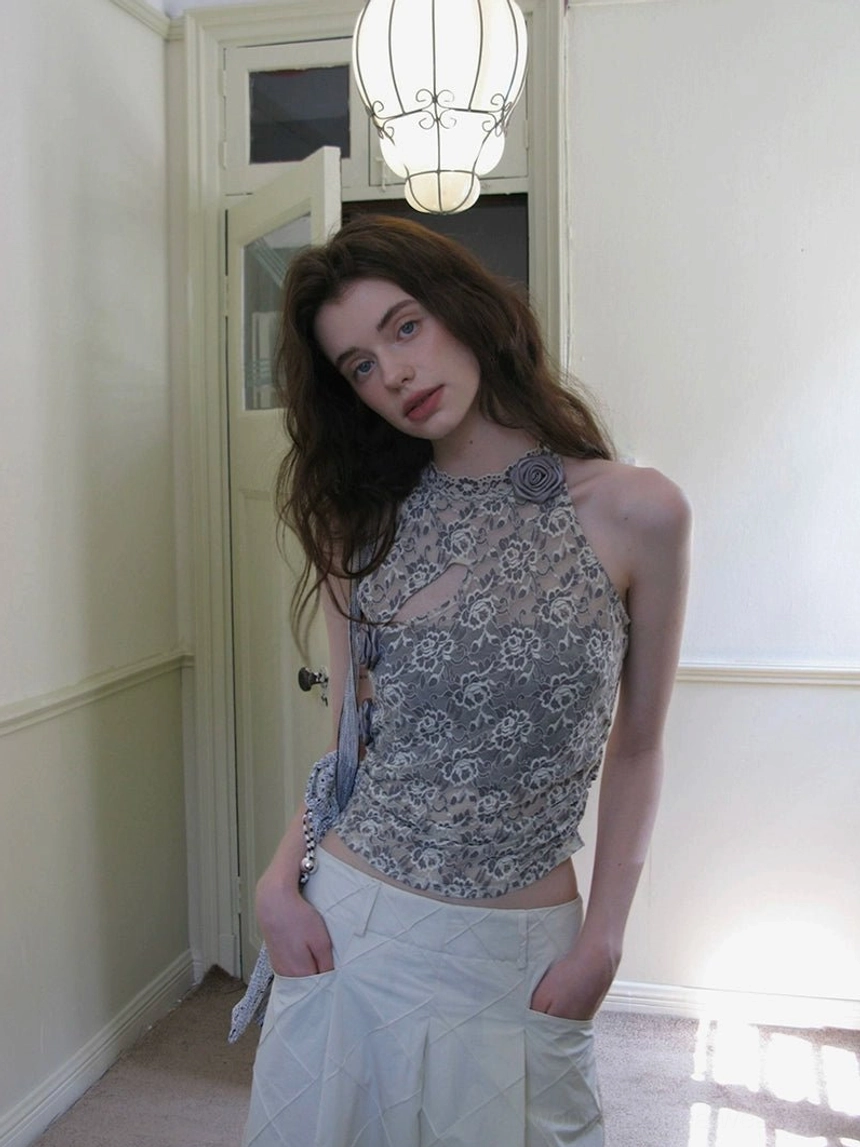 Model wearing a grey peony lace halter neck top with asymmetrical one shoulder silhouette and white pleated midi skirt in a retro indoor hallway