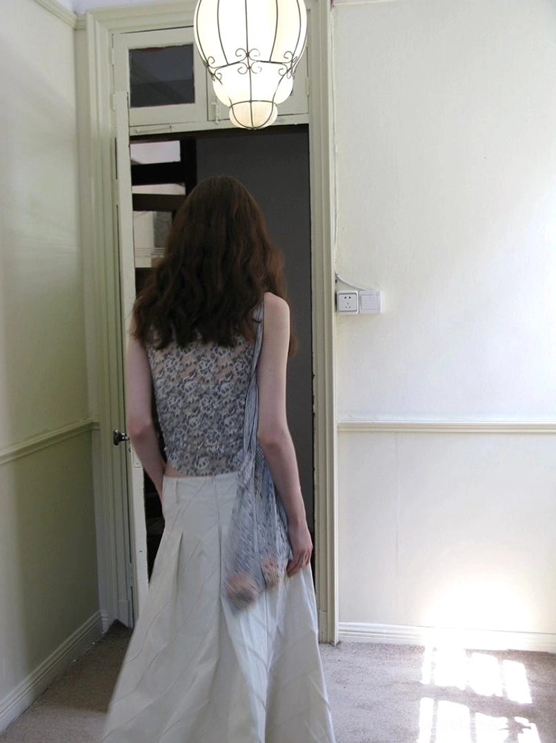 Back view of a grey peony lace halter neck camisole with backless styling and sheer side drape paired with a white long skirt