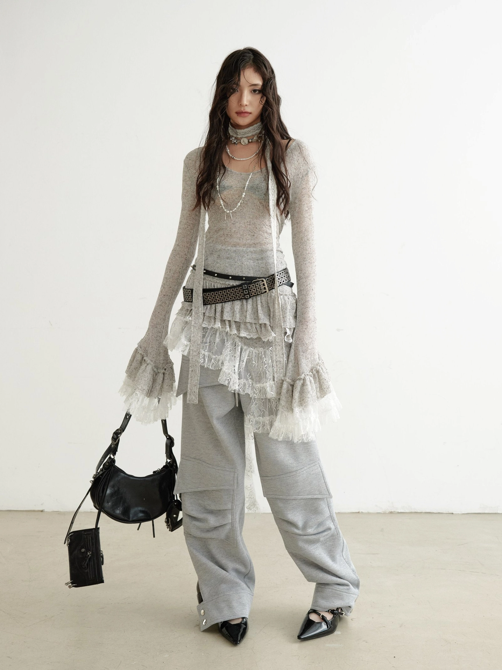 Full front view of grey semi-sheer lace knit dress with asymmetrical ruffle hem, layered necklaces, pointed heels, studded belt, and black crossbody bag