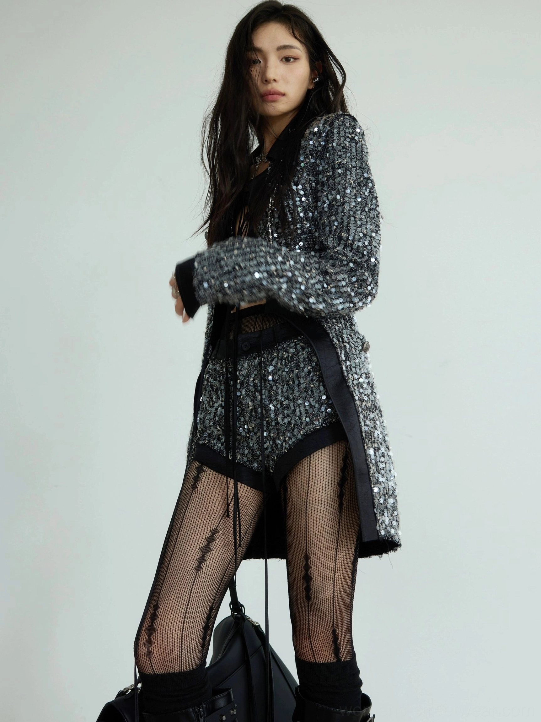Front three-quarter side view of a gunmetal sequin blazer and low-rise shorts matching set styled with striped fishnet tights and heeled boots
