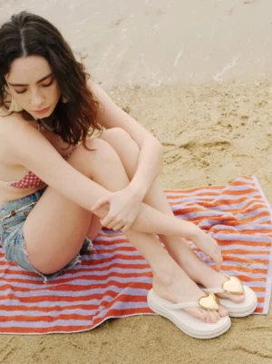 White gold heart platform flip-flops styled with denim shorts and a red check crop top on a sandy beach