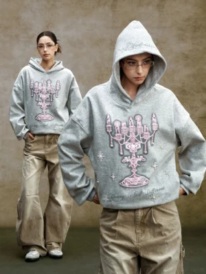 Front full-body view of a heather gray oversized chandelier graphic hoodie with pink embroidered candelabra, pearl-like embellishments, and two streetwear models wearing wide-leg khaki pants