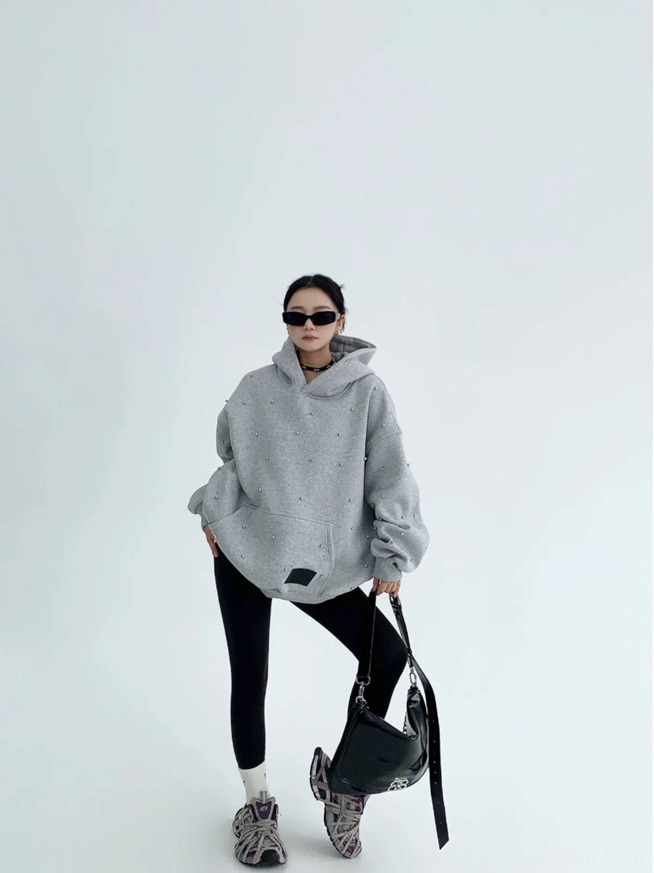 Full-body view of a woman wearing a heather gray oversized pearl-decorated hoodie with black leggings, sneakers, and a handbag
