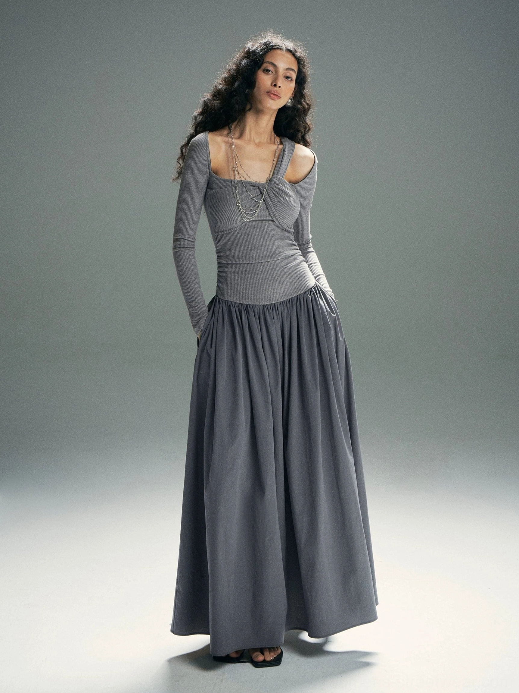 Heather gray twist neckline maxi dress with layered chain accent, asymmetrical cold shoulder, fitted bodice, and pleated floor-length skirt