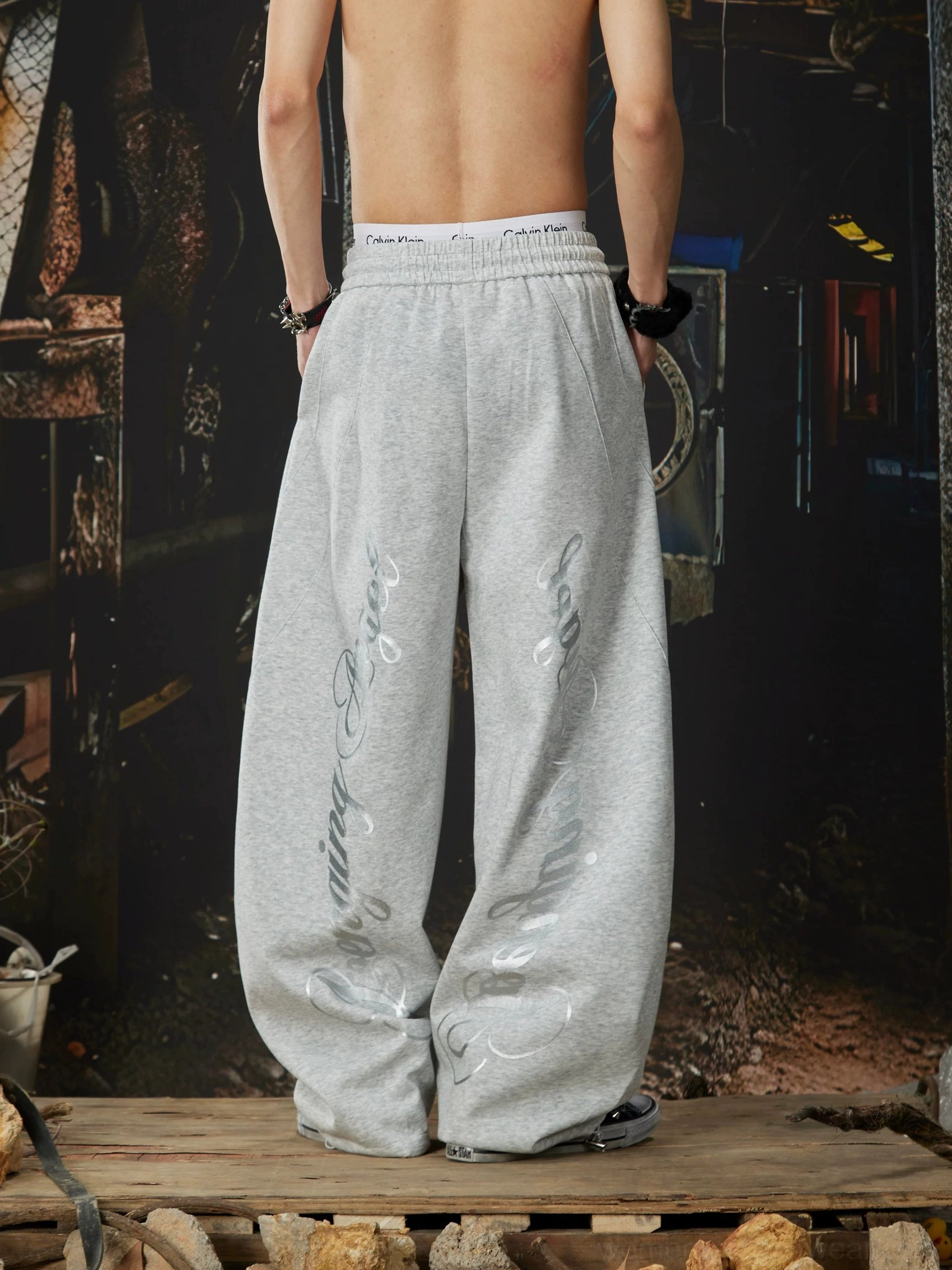 Heather grey gothic wide-leg sweatpants shown from the back with cursive script graphics on both outer legs and high-rise elastic waist