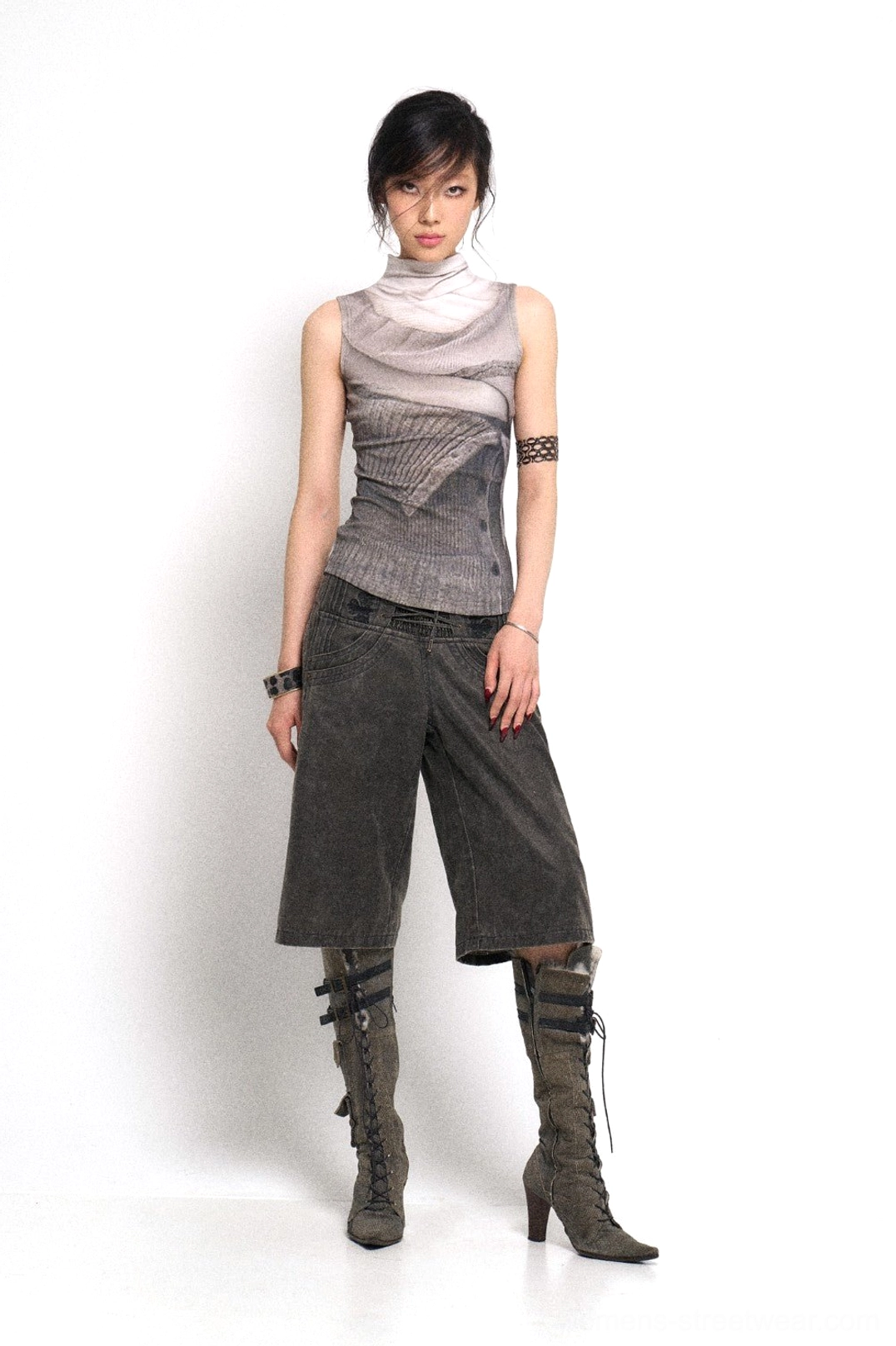 Full-body view of a model wearing high-rise washed denim culotte streetwear pants with a printed sleeveless top, lace-up heeled boots, and layered jewelry