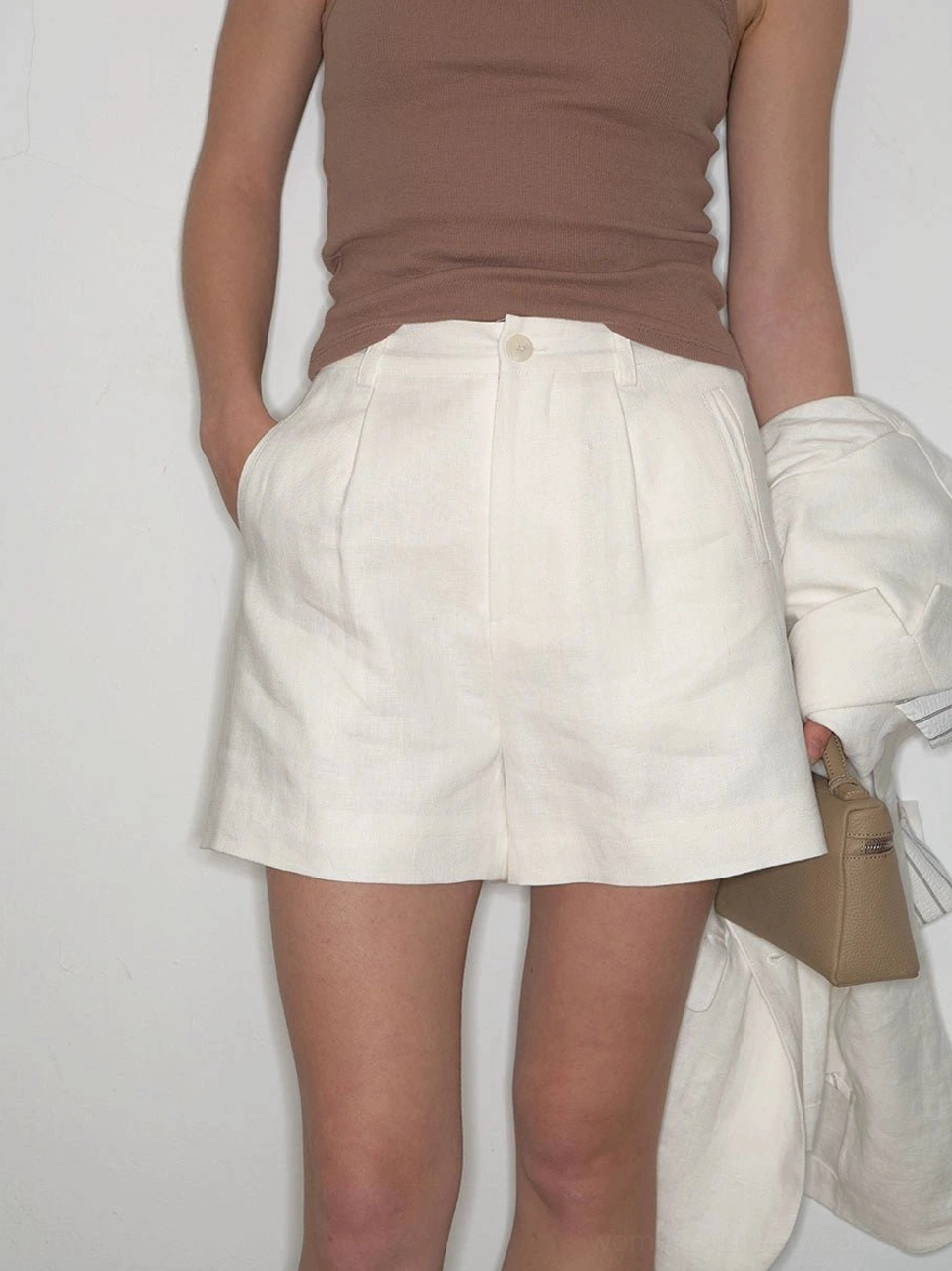 Detailed front view of a woman wearing high-waisted white linen pleated shorts with a brown tank top and blazer highlighting the textured fabric and relaxed straight-leg fit