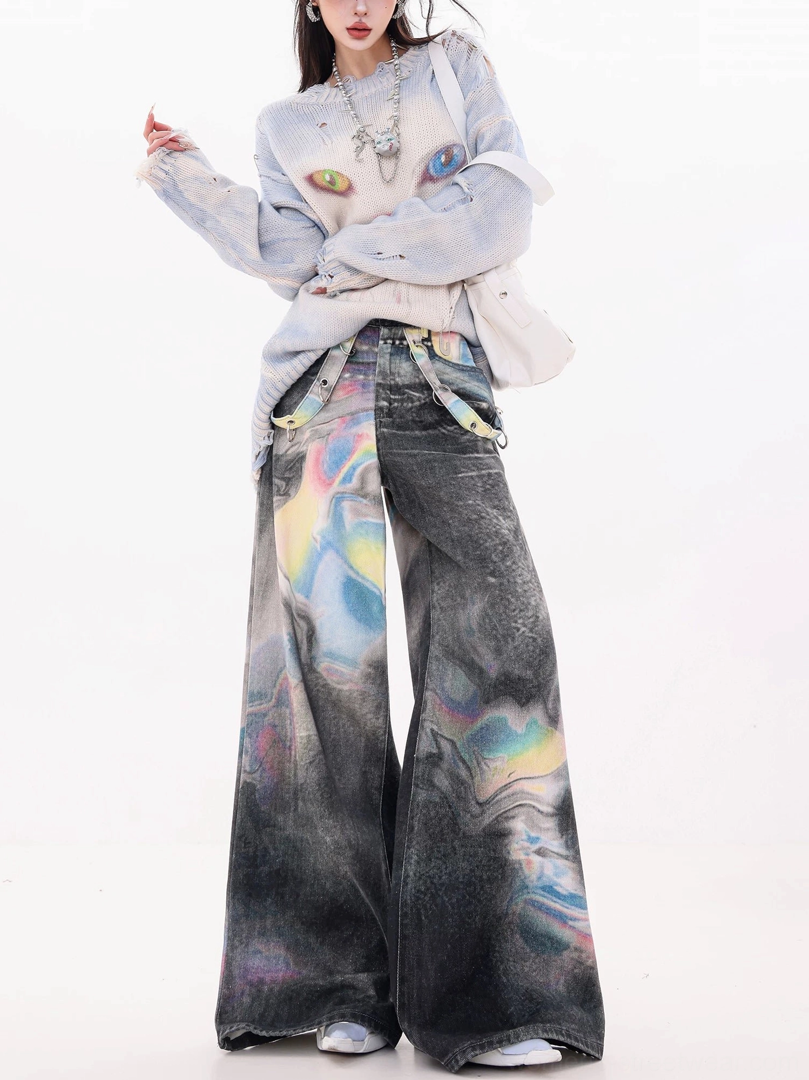 Full front view of women wearing holographic gradient wash ultra wide-leg jeans with a cropped graphic sweater, layered silver necklace, and white shoulder bag in a minimalist studio