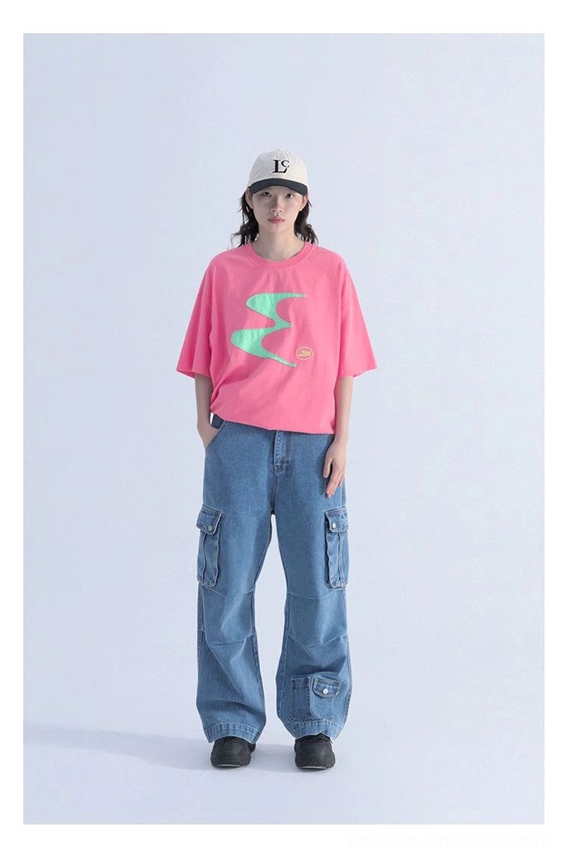 Full-body view of a woman wearing a hot pink oversized graphic tee with mint green wavy contrast patch design, blue wide-leg cargo jeans, and a baseball cap