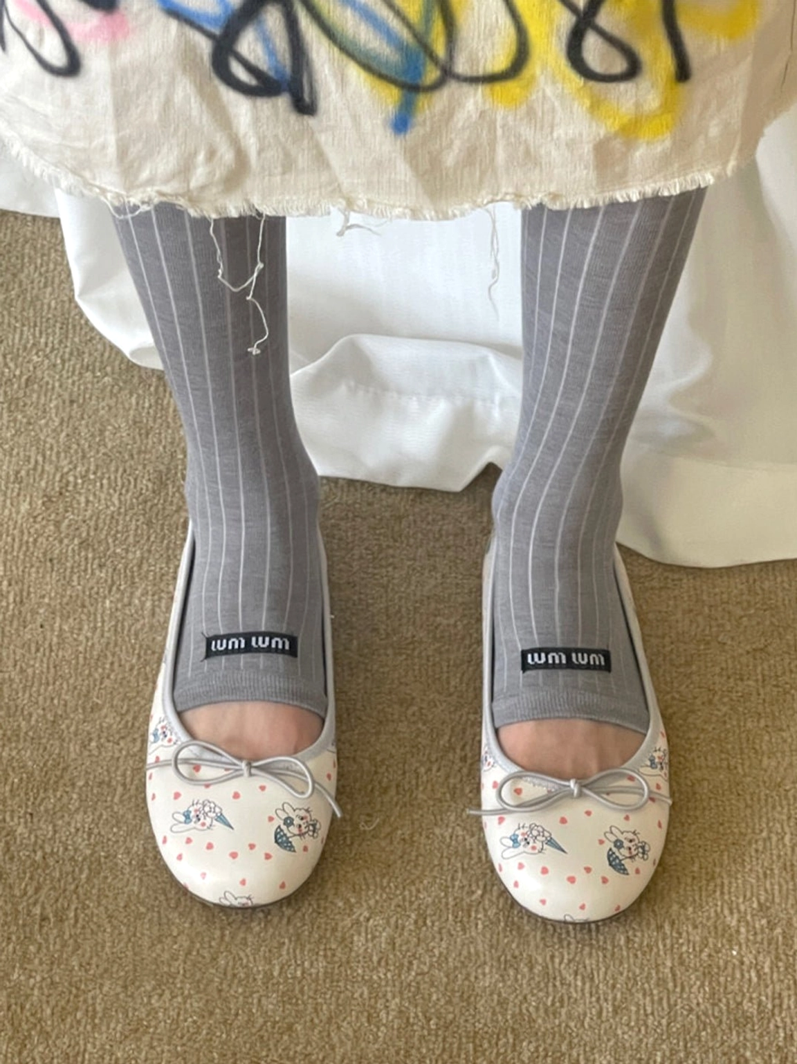 Top view of illustrated rabbit print ribbon ballet flats styled with gray socks and a graffiti print midi skirt