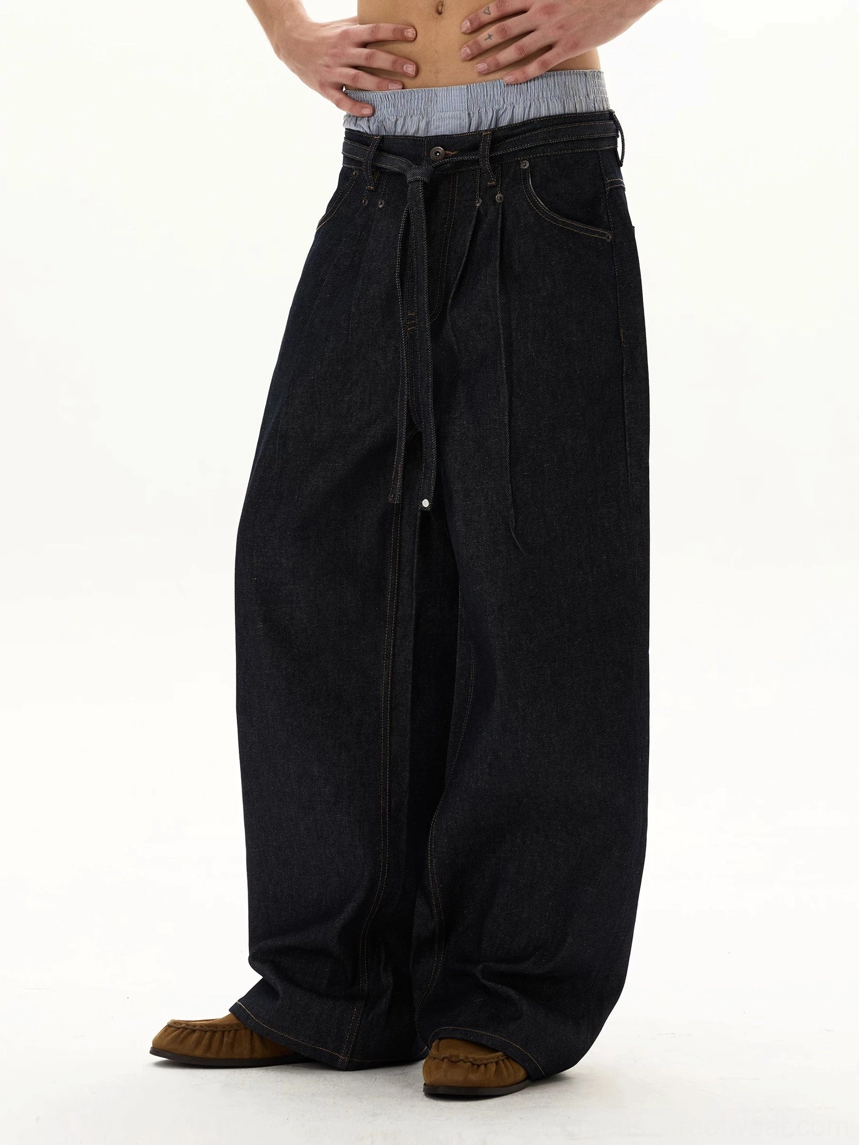 Three-quarter front view of dark indigo balloon jeans with layered waistband, adjustable drawstring tie, front pleats, and extra-wide legs