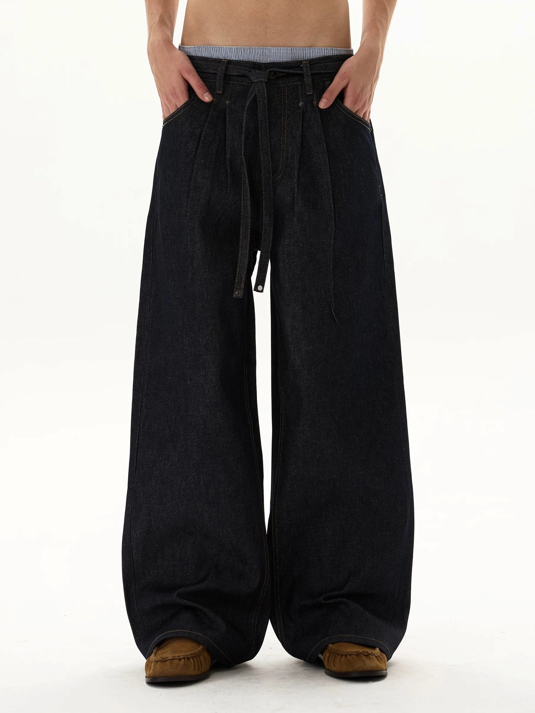 Front view of oversized indigo balloon jeans with ultra-wide puddle legs, adjustable tie waist, exposed waistband, and raw-style dark denim