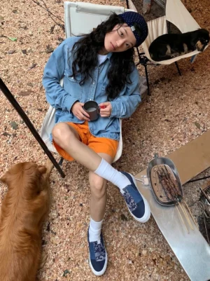Female model wearing indigo paisley patchwork slip-on sneakers with white crew socks at an outdoor gravel campsite streetwear setting