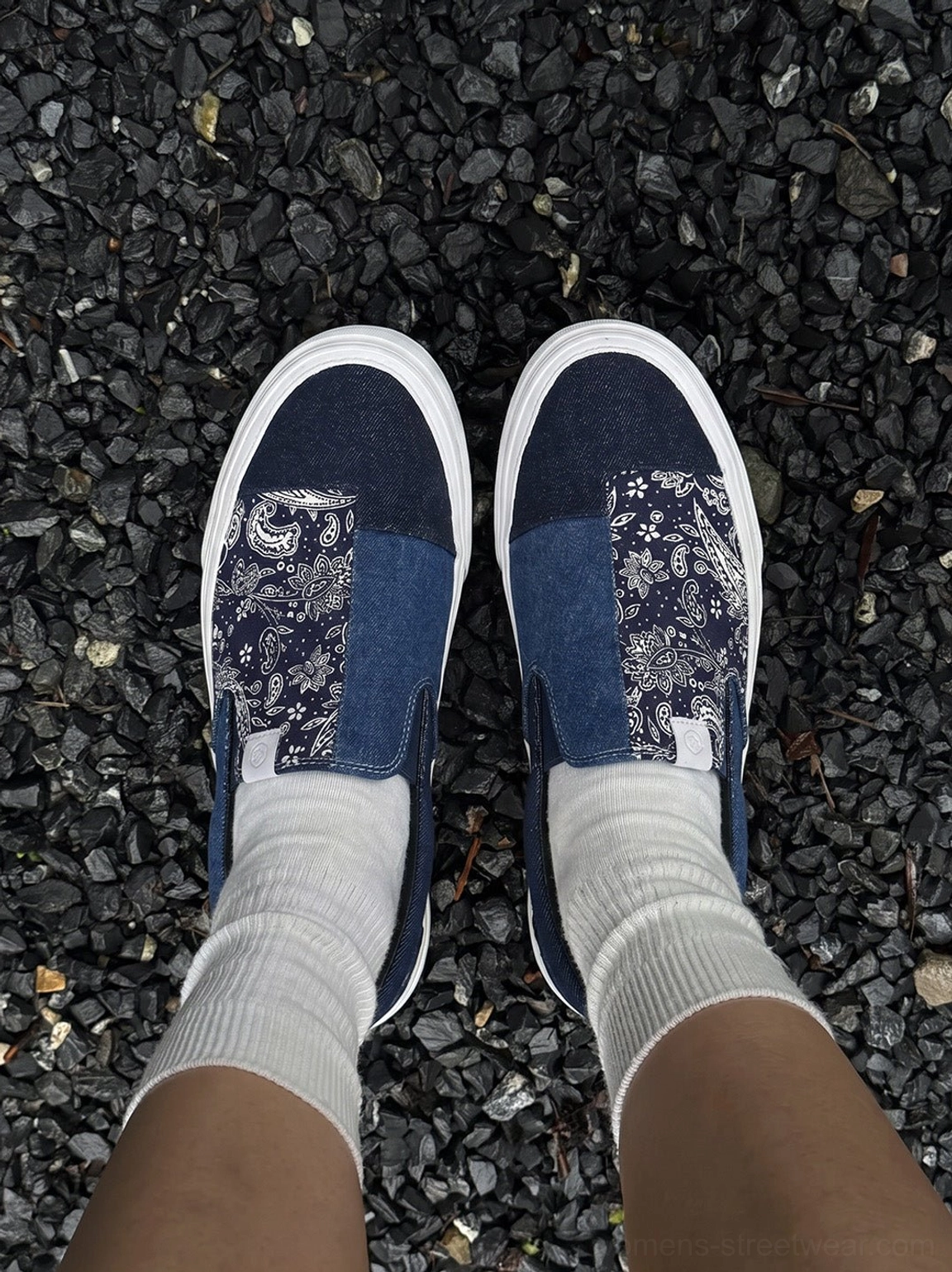 Top-down on-foot view of indigo paisley patchwork slip-on sneakers paired with white crew socks on black gravel
