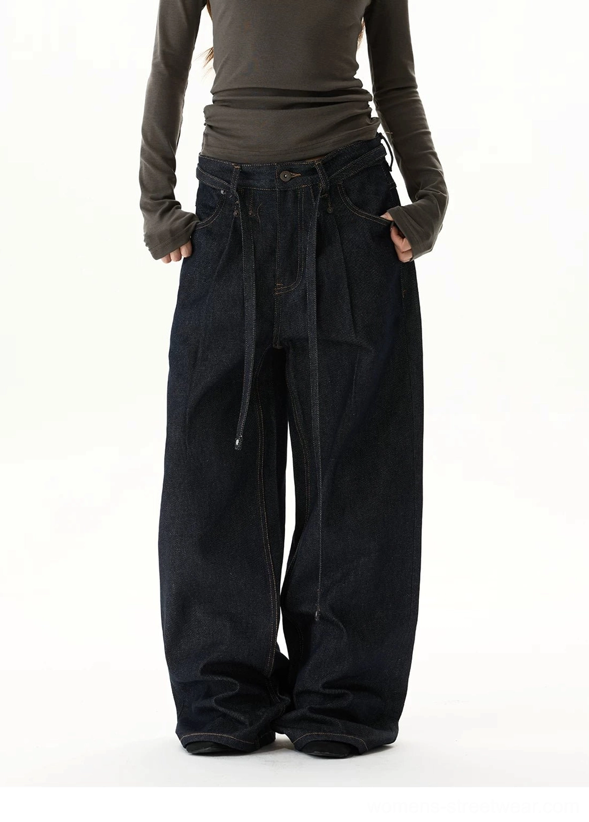 Three-quarter front view of indigo oversized wide-leg jeans with exposed layered waistband, tie detail, deep pockets, and relaxed streetwear styling