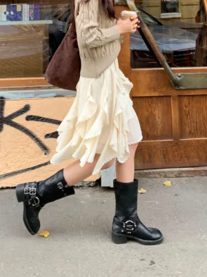Ivory asymmetric layered ruffle mini skirt styled with a khaki cable knit sweater and black buckle boots on an urban street