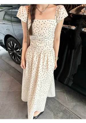 Side view of an ivory and black polka dot maxi dress with short flutter sleeves, fitted waist, and flowing floor length skirt styled for modern city streetwear