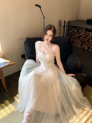 Ivory champagne floral embroidered tulle formal maxi dress shown seated, highlighting the ruched bust, dimensional floral bodice, and elegant one-shoulder neckline