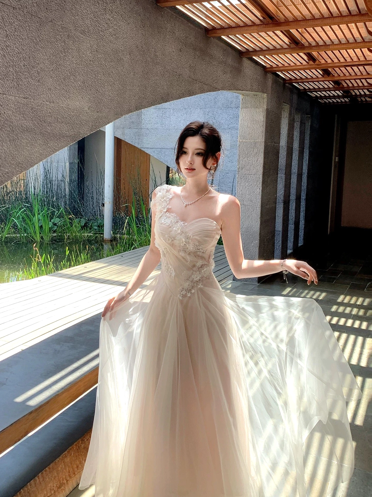 Side-front view of an ivory embroidered floral tulle maxi dress with a fitted waist and sweeping full-length skirt posed beside a waterside patio