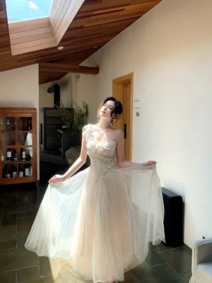 Full-body view of an ivory floral embroidered tulle one-shoulder maxi dress with a fitted bodice and flowing floor-length skirt in a sunlit villa