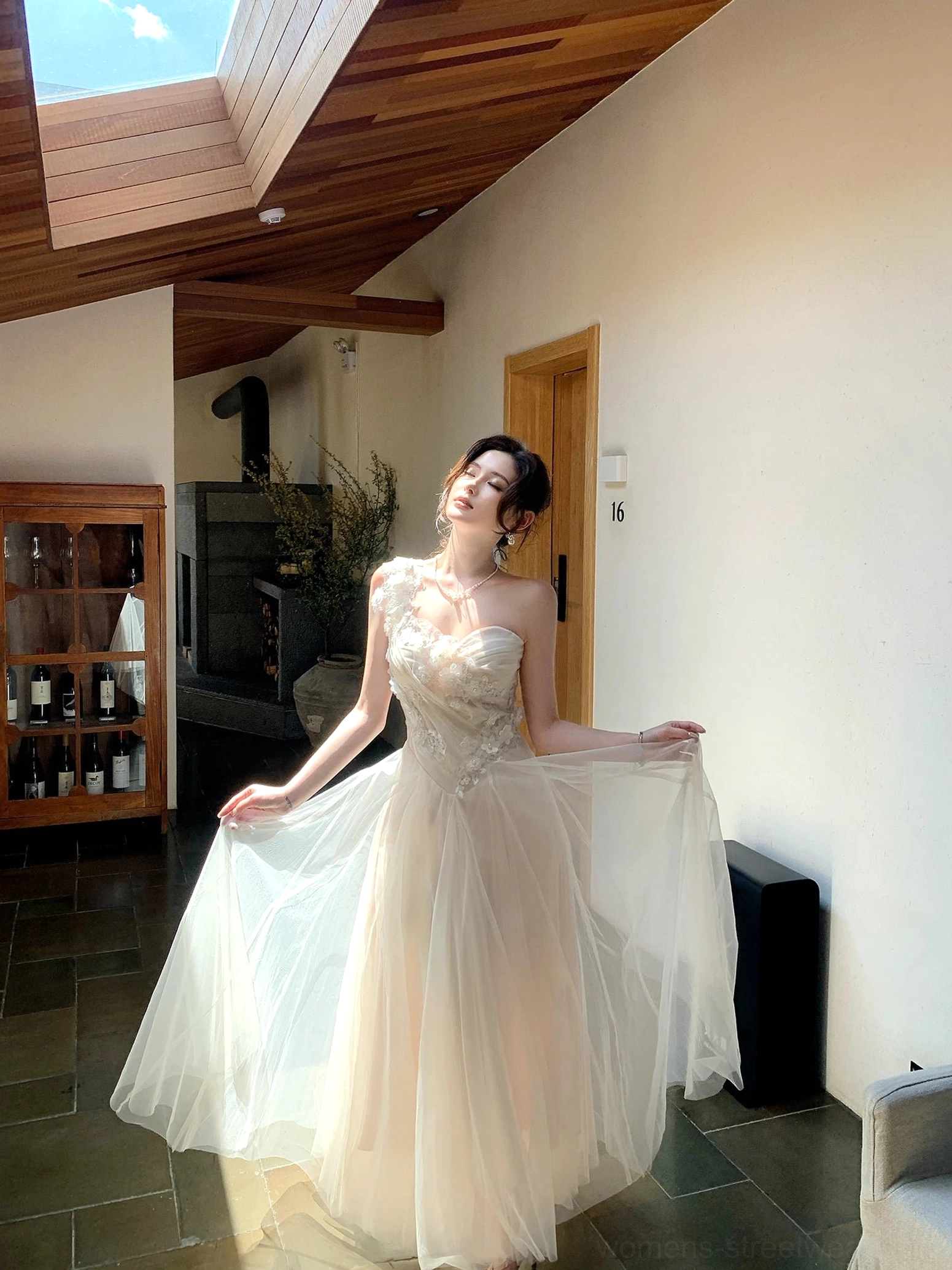Full-body view of an ivory floral embroidered tulle one-shoulder maxi dress with a fitted bodice and flowing floor-length skirt in a sunlit villa