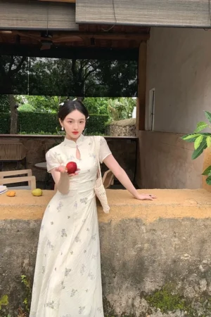 Front three quarter view of an ivory floral Mandarin collar cheongsam inspired maxi dress with keyhole neckline ruched waist short sleeves and flowing full length skirt styled with a woven shoulder bag