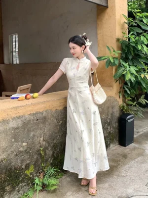 Side three quarter view of an off white floral Mandarin collar maxi dress with fitted waist short sleeves and floor length flowing silhouette styled with woven bag and flat sandals