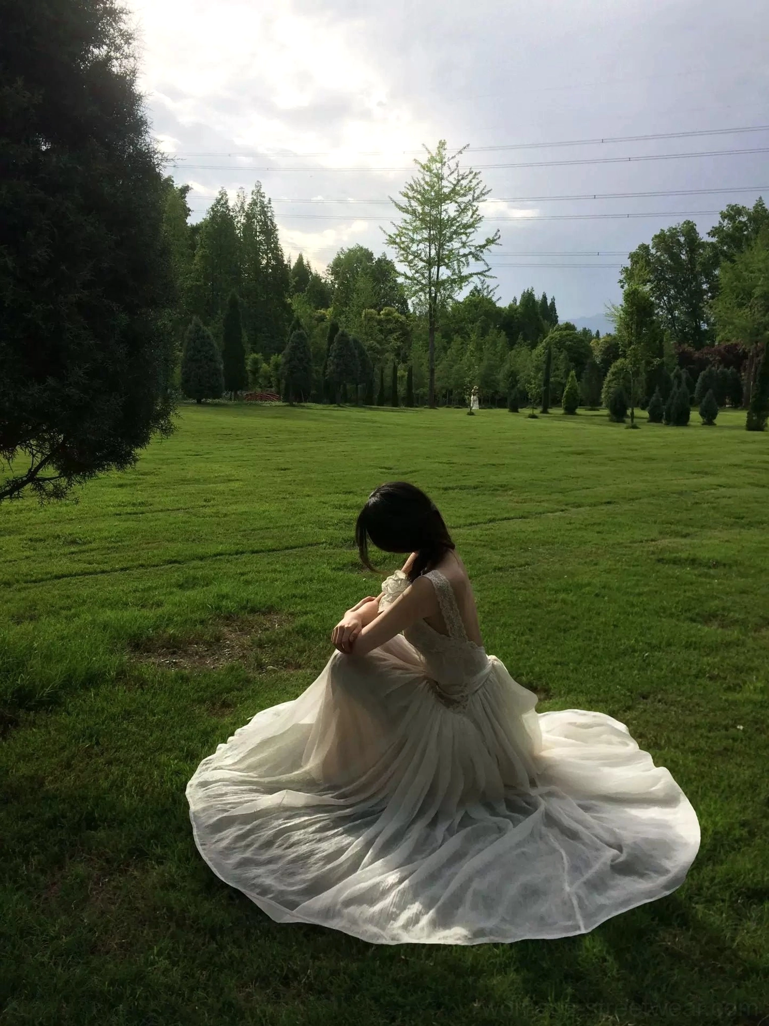 Seated view of an ivory lace open-back maxi dress with a low back, soft sheer flowing fabric, sweeping floor-length skirt, and romantic garden style