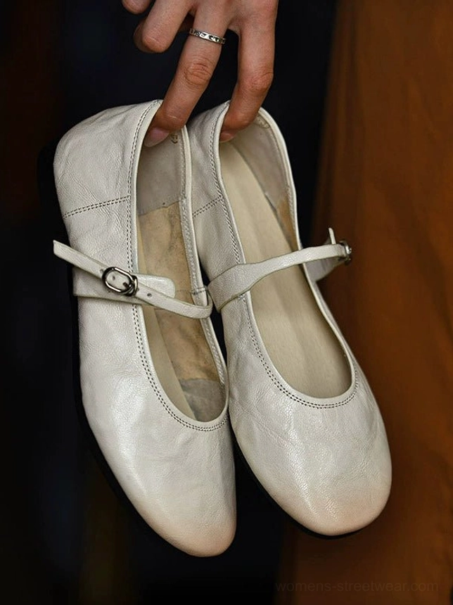 Pair of off white lambskin Mary Jane flats held from above, featuring soft textured leather, rounded toes, adjustable buckle straps, and stitched trim